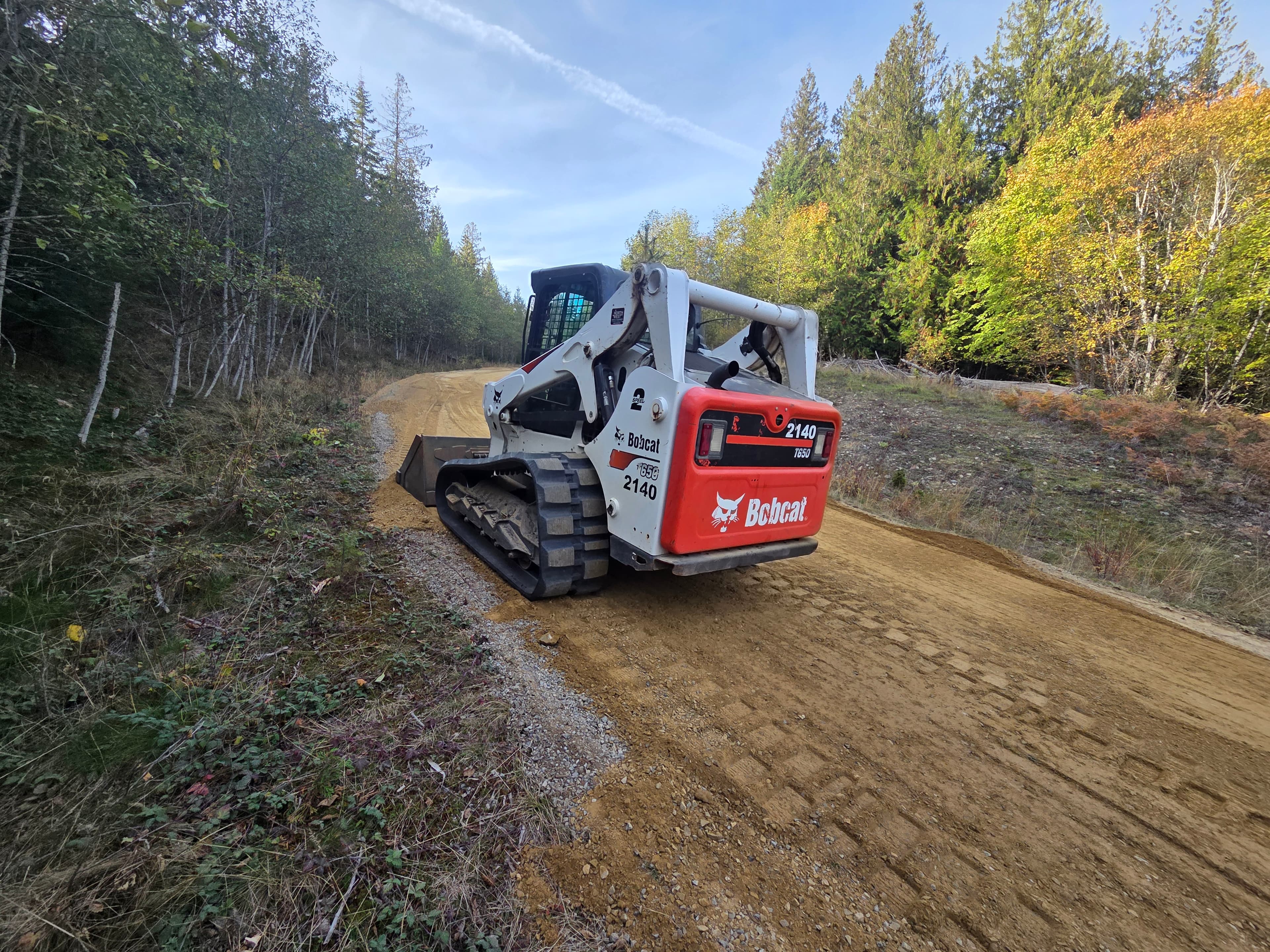 White Bobcat track loader grading a dirt road through a dense forest with autumn foliage.
