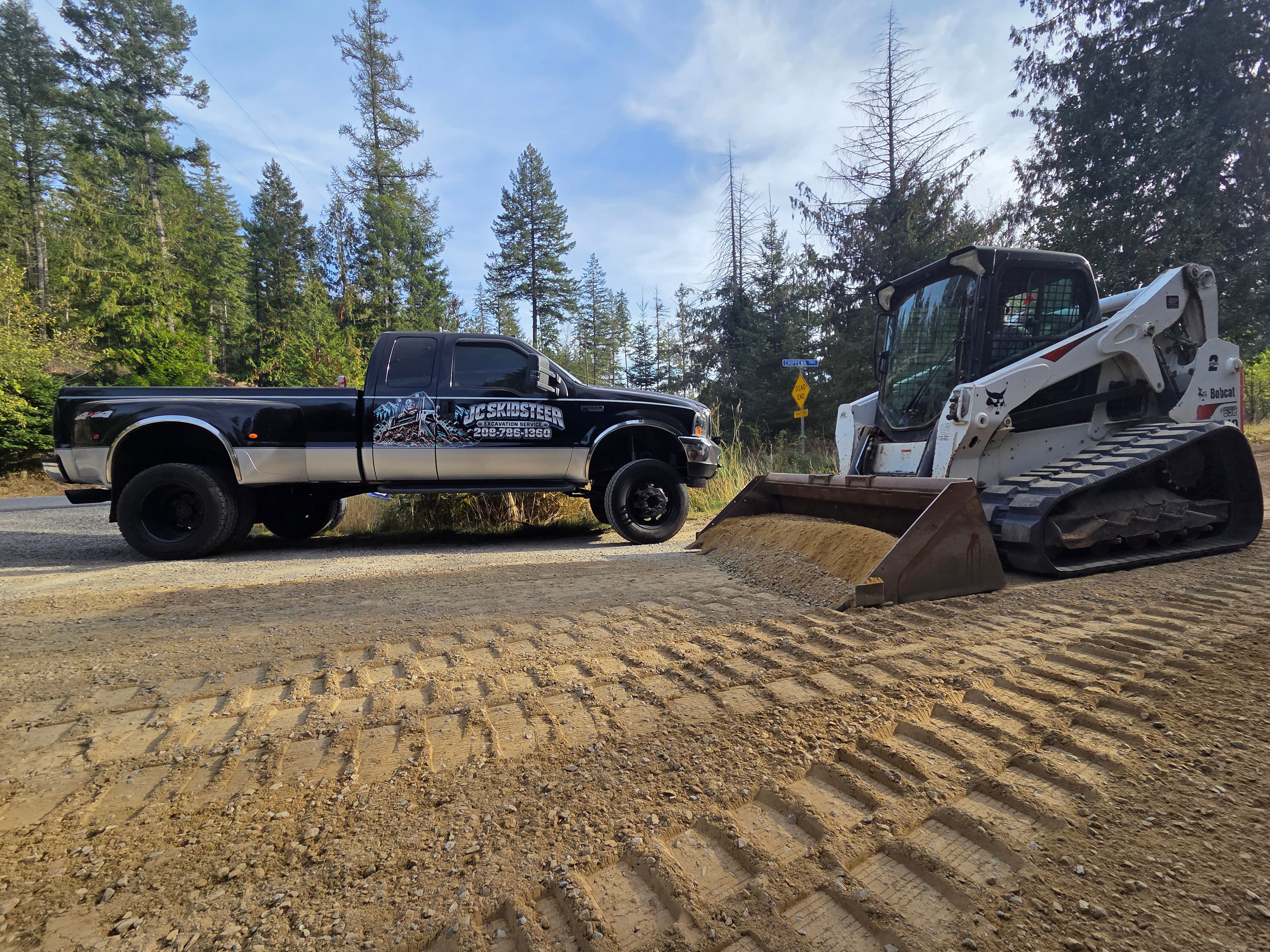 Black pickup truck and Bobcat skid steer loader on a dirt road in the woods.
