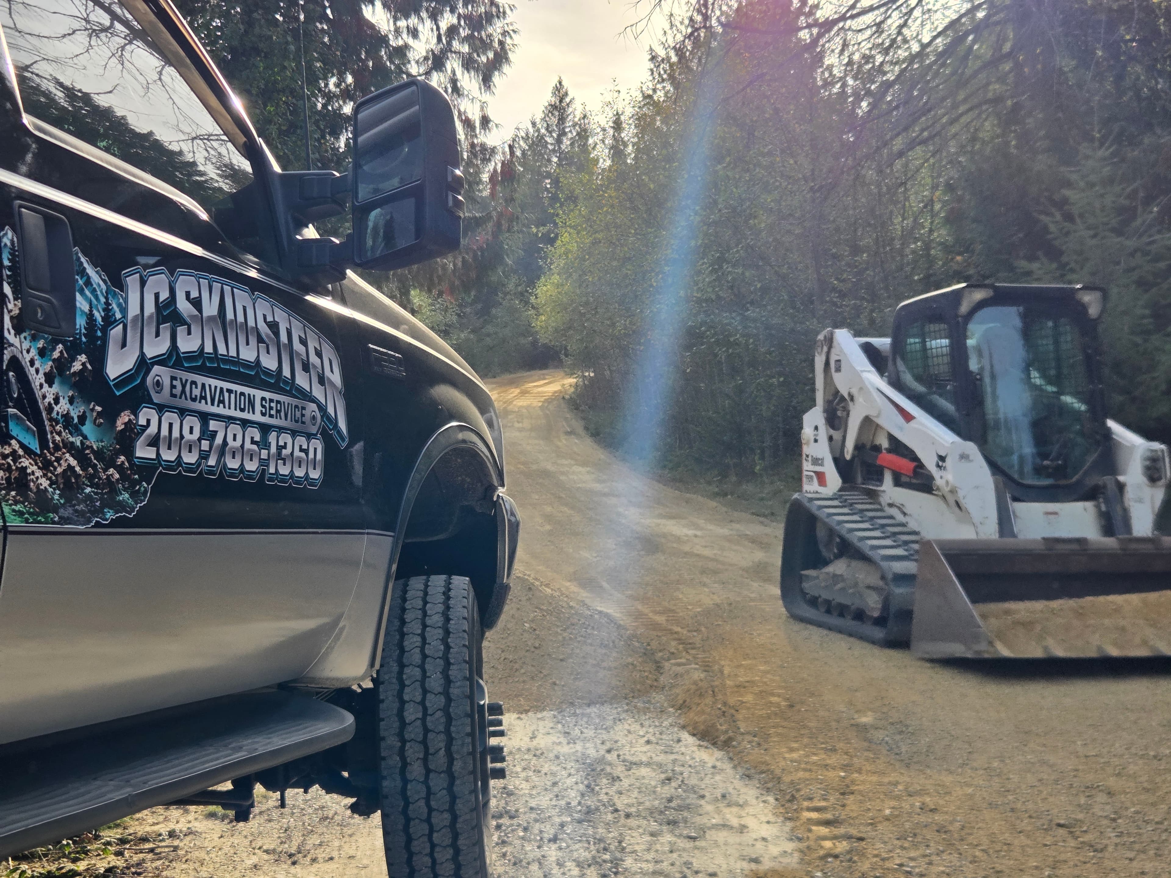Black excavation truck and white skid steer on a dirt road in a sunny forest.