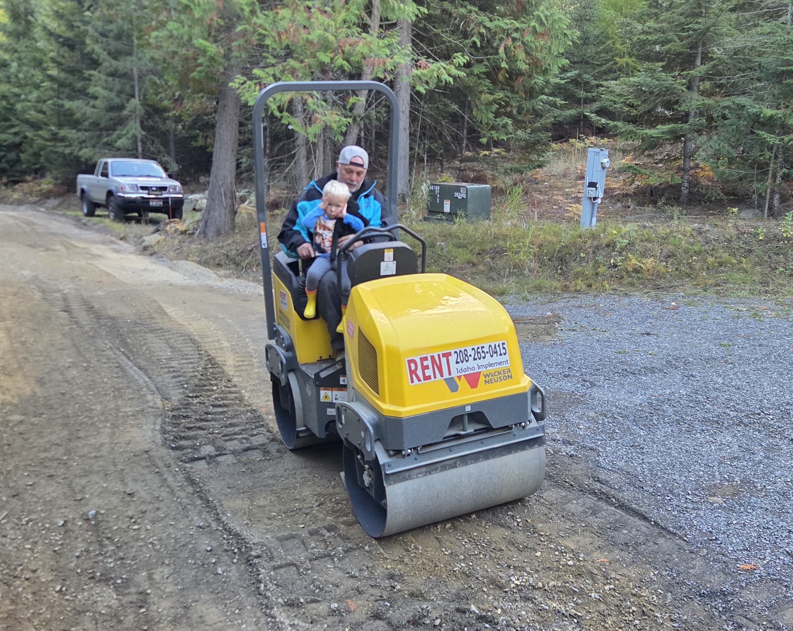 Man and young child sitting on a yellow rental steamroller on a dirt road.