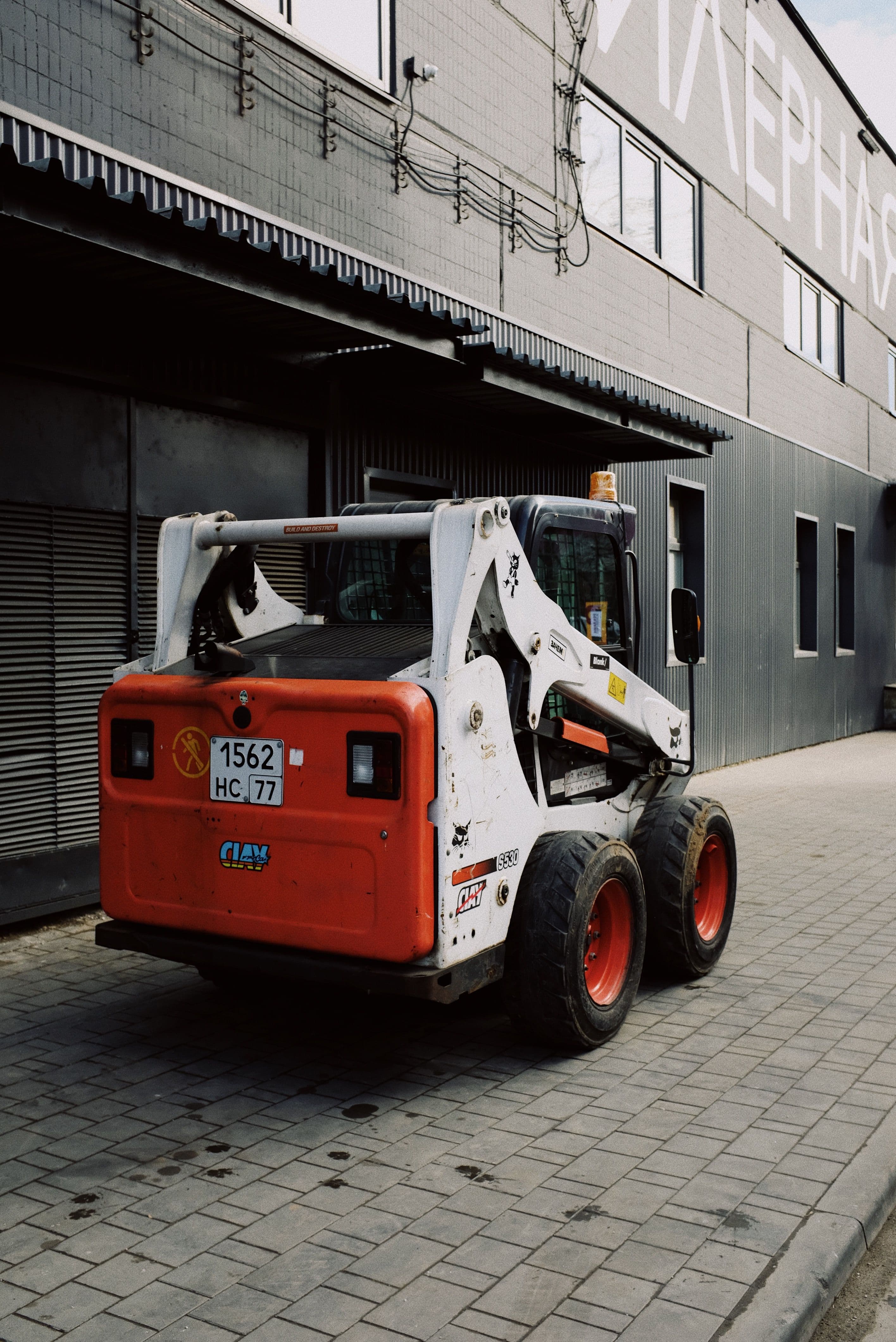 White and orange skid-steer loader parked on a paved sidewalk beside a grey building.